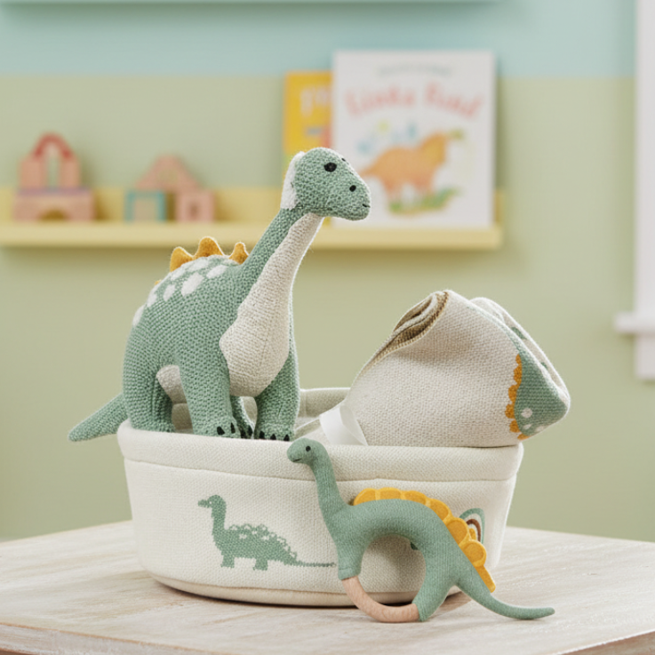 A set of plush dinosaur toys arranged in a round cream fabric basket with printed dinosaur silhouettes. The set includes a tall green-and-white long-neck dinosaur, a smaller green-and-white dinosaur with yellow spikes, a pale cream stuffed dinosaur head, and a green-and-white ring-shaped dinosaur toy.